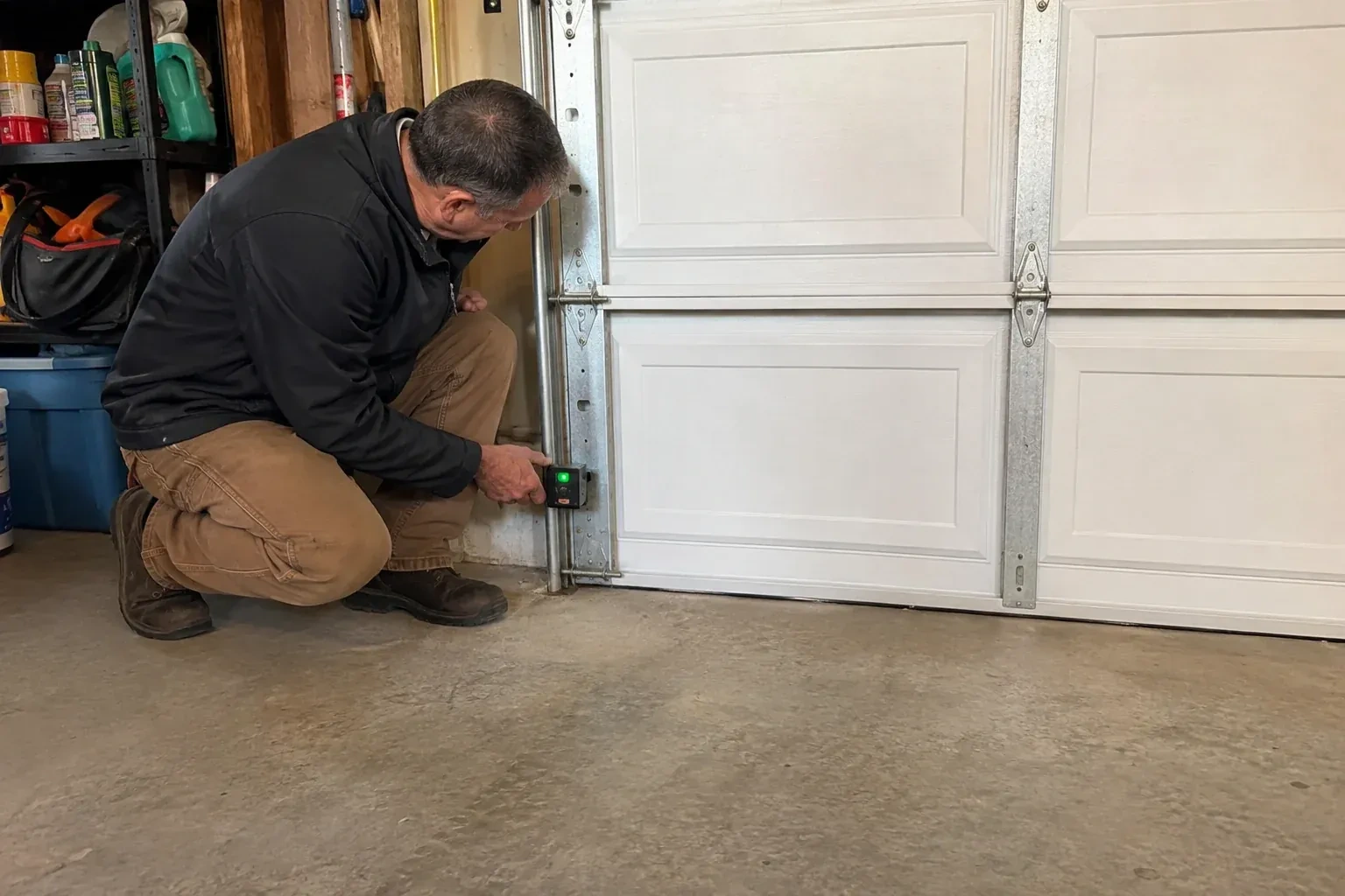 Technician kneeling beside a white paneled garage door, holding a small black box with a green light against the vertical track. — Photo-Eye Sensor Alignment or Replacement, Oakville