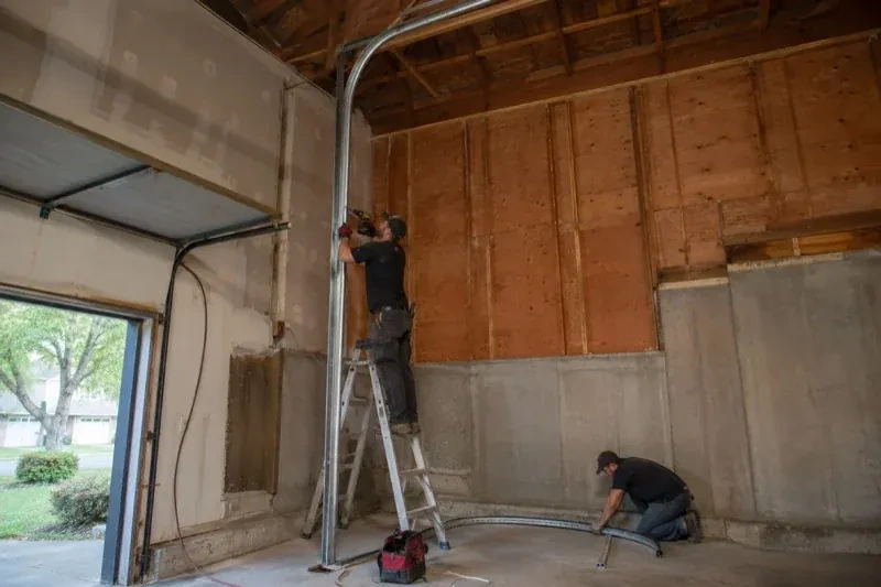 One person on a stepladder working beside a tall curved garage-door track while another kneels beside loose track sections in an unfinished garage. — High-Lift Garage Door Conversion, Oakville
